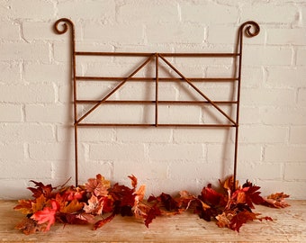May include: A rusty metal trellis with a geometric design, ideal for climbing plants. The trellis is standing in front of a white brick wall and is surrounded by a pile of autumn leaves.