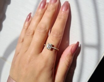 May include: A gold ring featuring a square clear stone surrounded by smaller stones. The ring is worn on a finger.