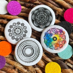 May include: Four white, circular buttons featuring mandala designs in black and white, and one with a rainbow dreamcatcher design. The buttons are on a woven, brown surface, with colourful felt circles.