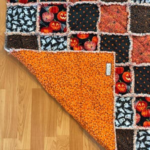 May include: A rag quilt featuring a patchwork of Halloween-themed fabric squares with pumpkins, ghosts, and spiderwebs in orange, black, and white. The quilt is folded back to reveal an orange backing fabric with small black polka dots.