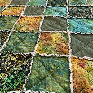 May include: A patchwork quilt featuring a range of green, brown and yellow fabrics. The quilt is made with squares of fabric that are sewn together with a white ruffle around the edges.