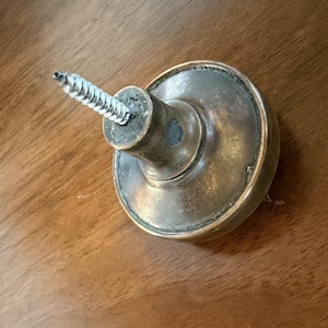Pode incluir: Puxador de porta de latão antigo com um parafuso prateado inserido. O puxador tem um design circular com um centro ligeiramente abaulado e uma mancha escura. O parafuso está parcialmente inserido no centro do puxador.