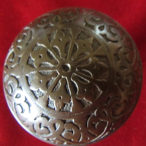 May include: A round, silver-coloured metal knob with an intricate, embossed design. The design features a central flower-like motif surrounded by smaller, repeating patterns.