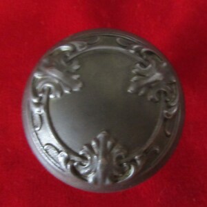 May include: A silver-toned metal trinket box with an ornate design featuring a circular centre and three stylised floral motifs.