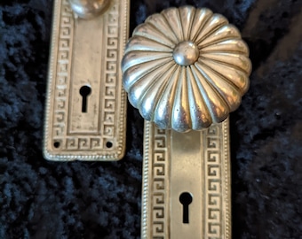 Può includere: Due pomelli per porte in ottone antico con un buco della serratura e un motivo decorativo a chiave greca. I pomelli hanno un design scanalato.