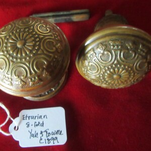 Peut inclure: Deux poignées de porte antiques en laiton avec des motifs floraux et volutes détaillés. Les boutons sont ronds et présentent une patine chaude et vieillie. Une petite étiquette indique "Etrurian 8-fold Yale & Towne £1899".