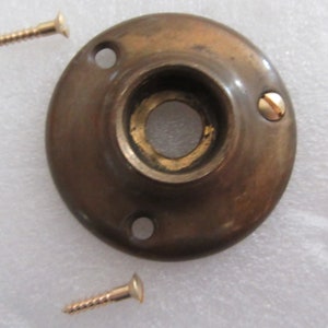 Op de afbeelding: Een messing deurknop-achterplaat met drie schroefgaten en twee schroeven. De achterplaat is rond en heeft een verhoogde rand.