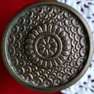 Op de afbeelding: Een rond, zilverkleurig metalen ornament met een gedetailleerd, ingewikkeld ontwerp. Het midden heeft een cirkelvormig patroon met een bloemvorm. De buitenste ring heeft een herhalend patroon van kleine, afgeronde vormen.