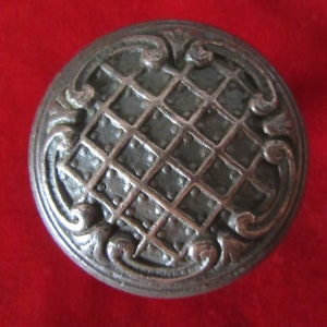 May include: A silver-toned door knob with a decorative, ornate design. The knob has a grid pattern in the center and a raised, floral design around the edge.