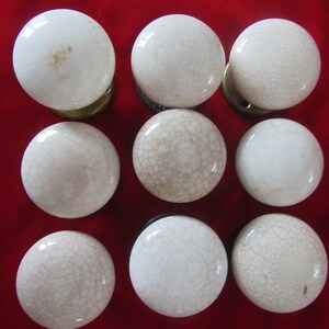 May include: Nine off-white ceramic door knobs with a crackle glaze pattern. The knobs are round with brass or black metal hardware. The knobs are arranged in a three-by-three grid on a red background.