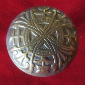 May include: A brass door knob with an intricate, geometric design. The knob is round and has a raised, circular pattern in the center.