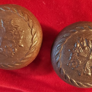 May include: Two brown, ceramic, round objects with a floral design in the center and a braided border. The objects are on a red background. The ceramic pieces have a rustic, handmade appearance.