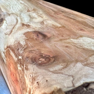 May include: Close-up of a wooden surface with a natural grain pattern. The wood features a mix of light and dark brown tones, with visible knots and swirling patterns. The surface appears smooth and polished.