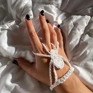 Puede incluir: Accesorio de mano blanco con forma de araña de ganchillo. La araña está conectada a una pulsera con una cadena. El texto "PDF PATTERN (EN) UNIQUE SIZE" es visible. El fondo es una superficie blanca y texturizada.