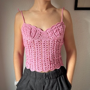 May include: A pink crochet top with delicate spaghetti straps and a scalloped neckline. The handmade top features an openwork design and a fitted silhouette. The top is paired with dark gray pants. This top is perfect for summer.