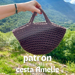 Puede incluir: Una cesta de ganchillo marrón con un asa trenzada se sostiene contra un telón de fondo de colinas verdes y cielo azul. La cesta tiene las palabras "patrón CROCHET cesta Amelie" en texto blanco.