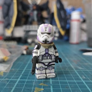 Può includere: Action figure di uno Stormtrooper bianco e viola con una pistola blaster nera. La figura ha un elmetto bianco con dettagli viola e una copertura per il collo color camoscio. La figura è su una superficie a griglia blu.