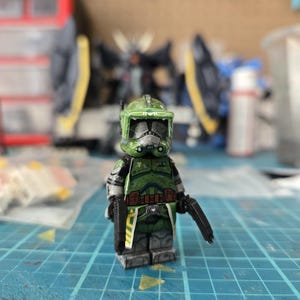 May include: A green and gray Lego minifigure of a clone trooper, holding two black blasters. The figure has a detailed helmet and armor, with white and brown accents. It stands on a blue grid surface.