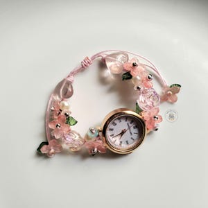 Puede incluir: Un reloj de pulsera con temática floral, con caja dorada y esfera blanca con números romanos negros. La correa de cordón rosa está adornada con flores rosas, cuentas y detalles transparentes. Un accesorio delicado y femenino.