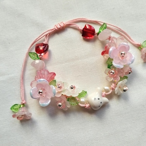 Puede incluir: Una pulsera rosa ajustable adornada con delicadas flores rosas y blancas, cuentas rojas y transparentes y un pequeño dije de conejito blanco. La pulsera está ensartada en un cordón rosa.