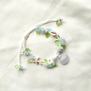 May include: A delicate bracelet featuring a white cord adorned with light blue flowers, green leaves, and small clear beads. The bracelet includes a seashell, a starfish, and a light purple seashell charm. The bracelet is on a white background.