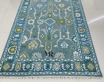 May include: A rectangular rug with a teal background and a detailed pattern in shades of blue, green, gray, and white. The rug features a central design with floral and geometric elements, framed by a border with repeating motifs. The rug has a fringe.