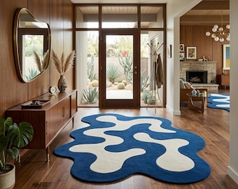 May include: An irregular, organic-shaped area rug featuring a bold, wavy pattern in deep blue and cream. The rug lies flat on a wooden floor, showcasing a fluid, abstract design that contrasts with the surrounding interior decor and furniture.
