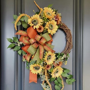 Peut inclure: Une couronne décorative composée de tournesols jaunes, de feuillage vert et de longues tiges florales jaune doré. Un grand nœud en ruban à carreaux orange et vert est fixé sur le côté, avec de longs pans de ruban tombant au centre de la couronne.