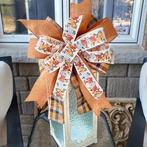 May include: A decorative fall bow in shades of orange, white, and yellow. The bow features a pumpkin-themed ribbon and is attached to a lantern. The lantern has a plaid pattern and is made of glass.