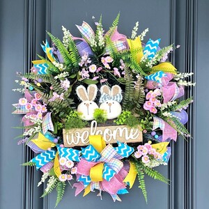 May include: A spring wreath with pink, yellow and blue ribbon bows, green ferns and pink flowers. Two bunny figurines are on a wooden sign that says "welcome".
