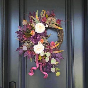 Peut inclure: Une couronne circulaire faite de brindilles brunes, ornée d'un bouquet de feuillage violet foncé, de deux grandes roses blanches, de petites fleurs violettes et orange, et de longues feuilles vertes, avec un ruban de velours violet suspendu.