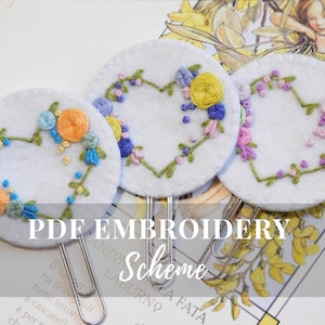 May include: Three white felt circles with embroidered floral heart designs in shades of pink, yellow, blue, and green. The circles are attached to silver binder clips. The text "PDF EMBROIDERY Scheme" is printed below the circles.