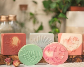 May include: Artisan soaps in various shapes and colors. The soaps feature botanical designs, including leaves and sunbursts. Colors include pink, green, and white. The soaps are displayed on a wooden board with dried flowers and a white cloth.