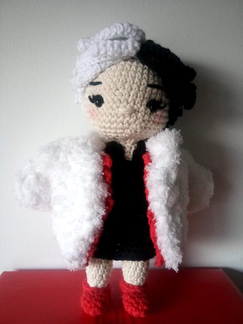 Puede incluir: Una mu&ntilde;eca de crochet con pelo blanco y negro, que lleva un abrigo de piel blanco con ribetes rojos y un vestido negro. La mu&ntilde;eca est&aacute; de pie sobre una superficie roja.