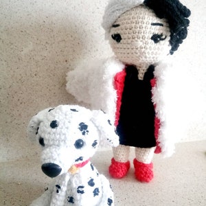 Puede incluir: Una mu&ntilde;eca de crochet con pelo blanco y negro que lleva un vestido rojo y un abrigo de piel blanco, de pie junto a un perro d&aacute;lmata de crochet.