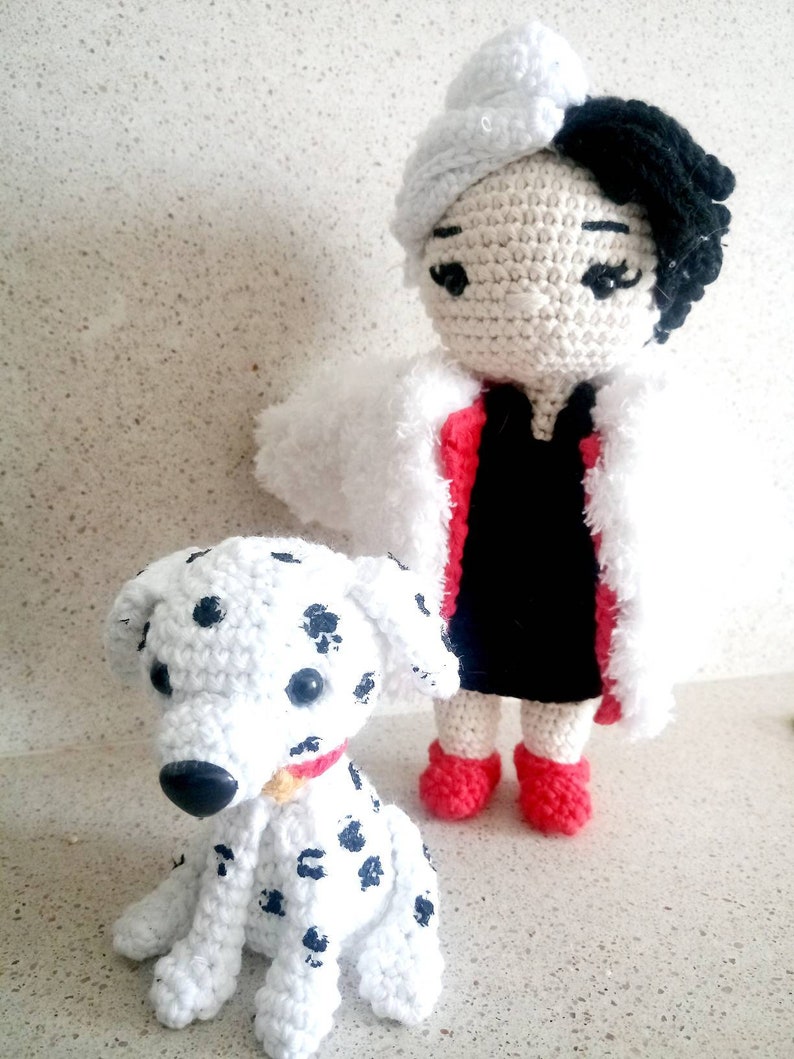 Puede incluir: Una mu&ntilde;eca de crochet con pelo blanco y negro que lleva un vestido rojo y un abrigo de piel blanco, de pie junto a un perro d&aacute;lmata de crochet.