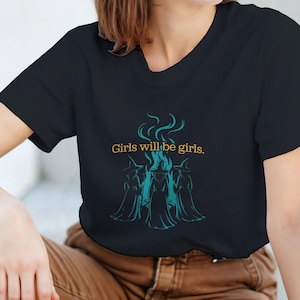 May include: Black t-shirt with a crew neck featuring the text "Girls will be girls" above a teal graphic of three figures around a flame. The shirt is made of a soft material.