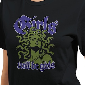 May include: A black short-sleeved t-shirt featuring a graphic print of a green Medusa figure with snakes for hair. Above the figure, the word Girls is printed in purple, and below the figure, the words will be girls are printed in purple.