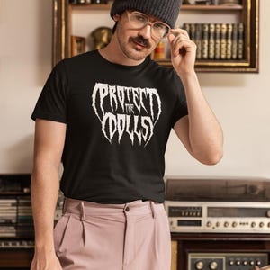 May include: A person wearing a black t-shirt with the text "Protect the Dolls" in white, a black beanie, and pink trousers. The person is standing in front of a bookshelf and a stereo.
