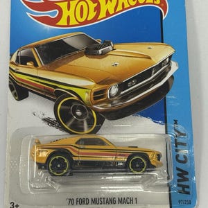 May include: A gold-colored '70 Ford Mustang Mach 1 Hot Wheels toy car on a blue and white card. The car has black and yellow stripes and black wheels. The card features the Hot Wheels logo and text that reads "Play Hot Wheels Showdown Game!" and "Guaranteed for Life."