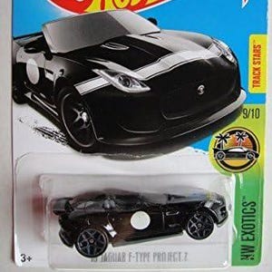 Puede incluir: Un coche de fundición negro Hot Wheels Jaguar F-Type Project 7 en una tarjeta azul y blanca. El coche tiene rayas blancas y un círculo blanco en el lateral. La tarjeta presenta el logotipo de Hot Wheels y el texto "HW Exotics".