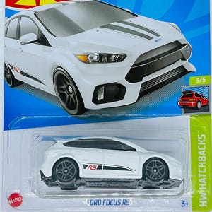 Könnte beinhalten: Ein weißes Ford Focus RS Hot Wheels Spielzeugauto mit schwarzen Rennstreifen und schwarzen Rädern. Die Verpackung enthält das Hot Wheels-Logo, den Text "HW Hatchbacks" und den Text "Ford Focus RS".