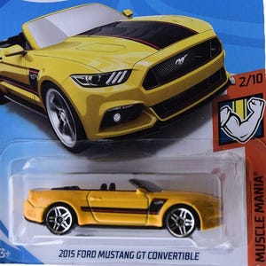 以下が含まれることがあります： 青とオレンジ色のホットウィールカードに、黄色の2015年フォードマスタングGTコンバーチブルのおもちゃの車。車には黒いストライプとアクセントがあります。カードには「Muscle Mania」と「2/10」の文字が含まれています。