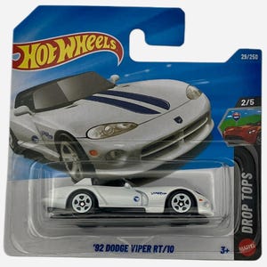 Puede incluir: Un modelo de fundición a presión Hot Wheels blanco de un '92 Dodge Viper RT/10. El coche de juguete tiene rayas azules y está empaquetado en una tarjeta azul y blanca. El embalaje incluye el logotipo de Hot Wheels y el texto "DROP TOPS".