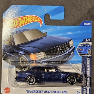May include: A blue Hot Wheels die-cast model of a '89 Mercedes-Benz 560 SEC AMG. The car is displayed in its original packaging, with the Hot Wheels logo and the text "'89 MERCEDES-BENZ 560 SEC AMG" visible. The packaging also includes the numbers 186/250 and 2/5.