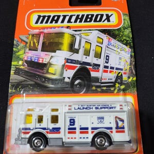 Peut inclure: Un camion de pompiers jouet Matchbox dans un emballage plastique transparent. Le camion blanc a des détails rouges et bleus, le numéro 9 et le texte "LAUNCH SUPPORT". L'emballage est orange et porte le logo Matchbox et les mots "HAZARD SQUAD".
