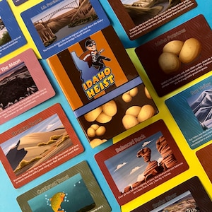May include: A board game titled "Idaho Heist" with cards featuring Idaho landmarks and illustrations. The game box is brown and orange, surrounded by cards with images of the Perrine Bridge, Idaho potatoes, and other scenic locations.