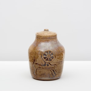May include: A brown ceramic jar with a lid. The jar features an embossed design of a spinning wheel. The jar has a rounded body and a slightly darker brown color at the base. The lid is a lighter shade of brown.