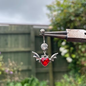 Może przedstawiać: Srebrny kolczyk do pępka z czerwoną zawieszką w kształcie serca i małymi srebrnymi skrzydłami. Pierścionek ma dwa przezroczyste, okrągłe klejnoty. Zawieszka jest zawieszona na srebrnym pręcie z małym, okrągłym klejnotem.