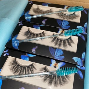 Op de afbeelding: Drie zwarte dozen met vlinderontwerpen bevatten individuele sets met valse wimpers en een blauwe wimperapplicator.